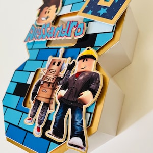 May include: A blue and gold 3D number 8 cake topper with a Roblox theme. The number is decorated with blue and white brick pattern, a robot, a Roblox character, and the name "Alessandro".