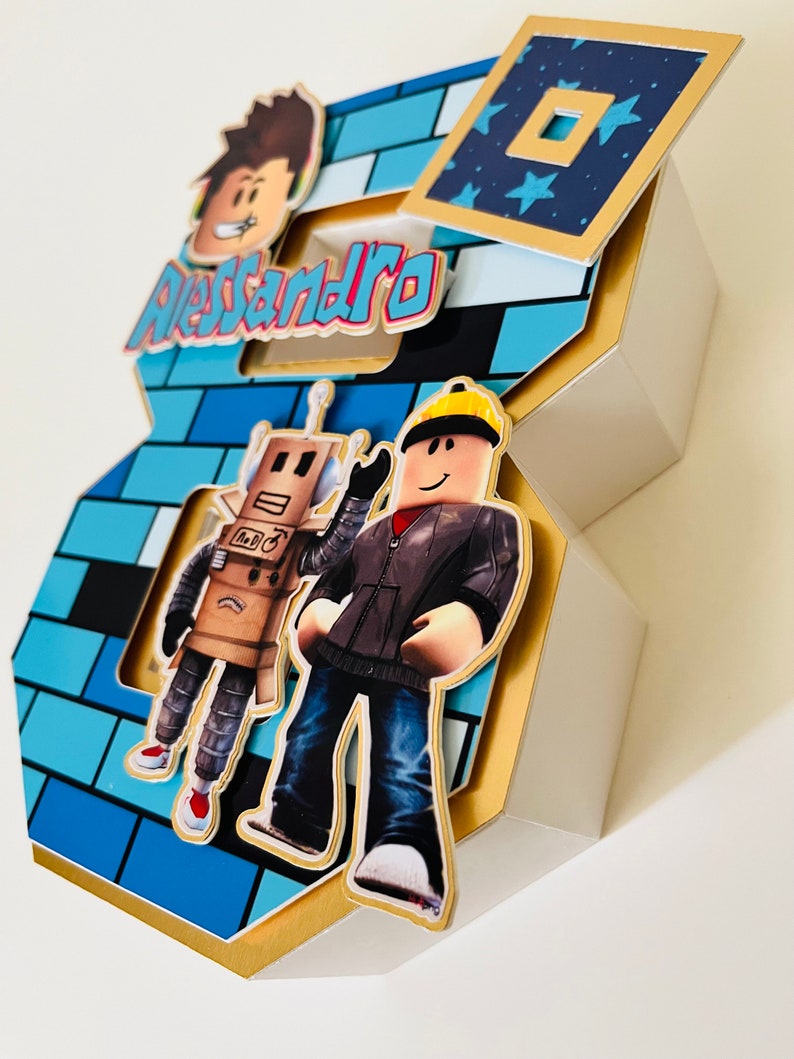 May include: A blue and gold 3D number 8 cake topper with a Roblox theme. The number is decorated with blue and white brick pattern, a robot, a Roblox character, and the name "Alessandro".