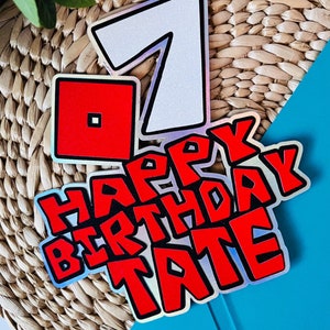 May include: A red and white cake topper with the number 7, a red square, and the words "Happy Birthday Tate" in a red and black font. The topper is on a blue and brown background.