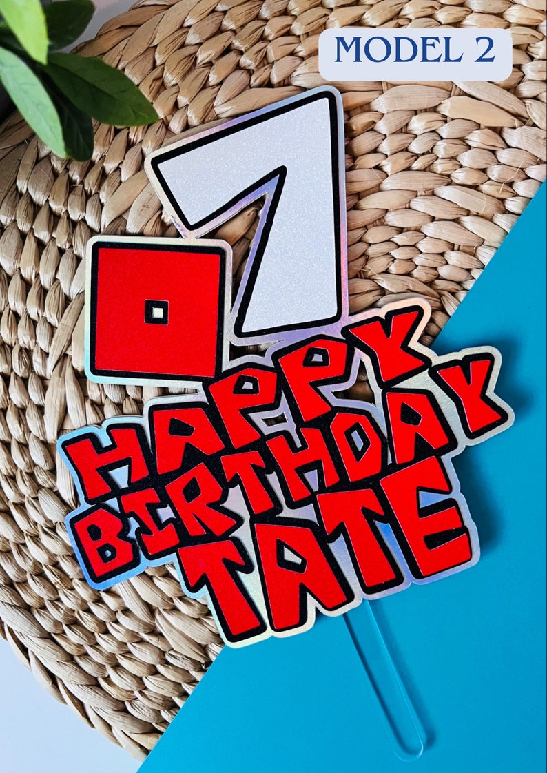 May include: A red and white cake topper with the number 7, a red square, and the words "Happy Birthday Tate" in a red and black font. The topper is on a blue and brown background.