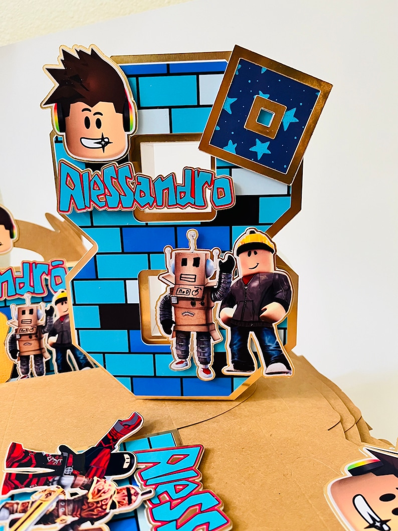 May include: A blue and gold Roblox themed cake topper with the number 8, a brick pattern, and the name Alessandro. The topper features Roblox characters, including a robot and a figure wearing a black jacket.