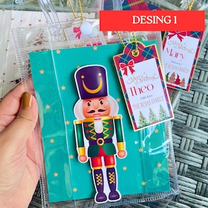 May include: A Christmas gift tag with a red and green plaid background and gold trim. The tag features a festive design with a Christmas tree and the text "Merry Christmas Theo With love The Rojas Family".
