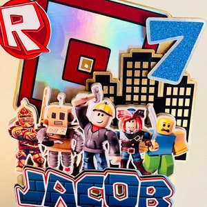 May include: A Roblox themed cake topper with a red and white "R" logo, a blue number 7, and a cityscape. There are five Roblox characters, including a knight, a robot, a builder, a pirate, and a yellow character. The name "JACOB" is written in blue letters on a brick background.