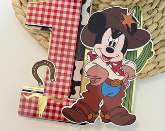 May include: A red and white checkered number one cake topper with a cartoon cowboy Mickey Mouse wearing a brown hat with a gold star. Mickey Mouse is holding a green cactus and has a red bandana around his neck.