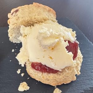 Può includere: Un scone appena sfornato, tagliato e ricoperto da un generoso strato di panna rappresa e una cucchiaiata di marmellata rossa. Lo scone è marrone chiaro e poggia su una superficie grigio scuro, una delizia classica.