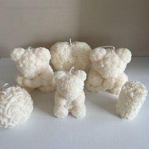 Könnte beinhalten: Ein Set aus sechs weißen Rosenkerzen in Form eines Herzens, eines Bären und einer runden Kugel. Die Kerzen sind aus weißem Wachs und haben ein zartes Rosenmuster.