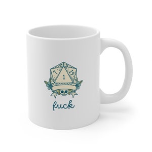 May include: White ceramic mug with a tattoo-style design of a D20 die, a skull, and floral accents. The number 1 is displayed on the die. The word "fuck" is written below the design. The mug has a curved handle.
