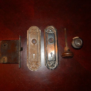May include: Antique door hardware set with a silver finish. The set includes a door knob, a door plate, a latch, and a spindle. The door knob is round with a keyhole and a decorative design. The door plate is rectangular with a keyhole and a decorative design. The latch is rectangular with a keyhole. The spindle is a long, thin metal rod.
