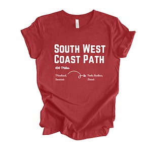 Puede incluir: Camiseta roja con texto blanco que dice "South West Coast Path" y "630 Miles". El texto también incluye "Minehead, Somerset" y "Poole Harbour, Dorset" con una línea y una flecha que apunta de izquierda a derecha.