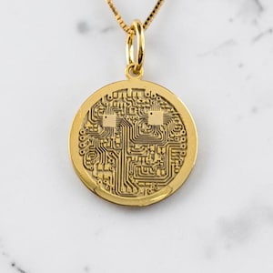 Pode incluir: Um colar com pingente dourado com um berloque circular com um intrincado design de placa de circuito. O pingente pende de uma delicada corrente dourada. O colar é exibido em um fundo de mármore branco.