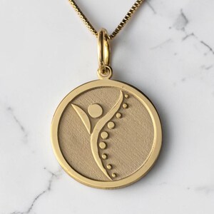 May include: A gold-colored pendant necklace featuring a circular pendant with a stylized figure and a curved line of dots. The pendant hangs from a delicate gold chain, set against a marble background. The design is simple and elegant.