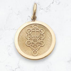 May include: A circular gold pendant featuring an engraved Tree of Life geometric design. The pendant has a smooth, raised outer rim and a textured central field. A small, vertical gold bail is attached to the top for hanging the piece.