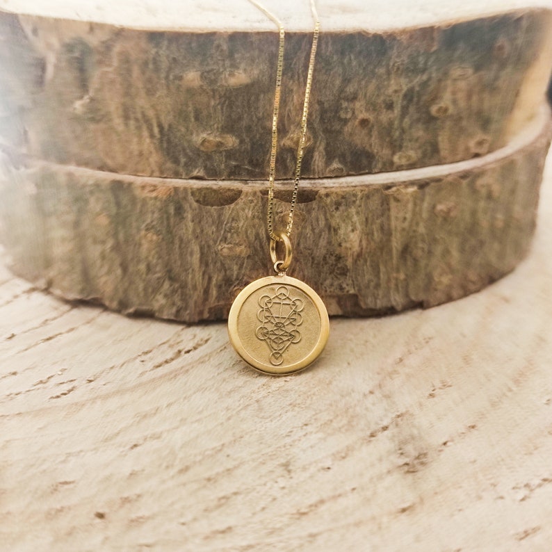 May include: A gold-toned circular pendant necklace featuring a detailed geometric Tree of Life engraving on its face. The pendant is suspended from a delicate gold-toned box chain, showcasing a polished metallic finish and a centred, intricate design.