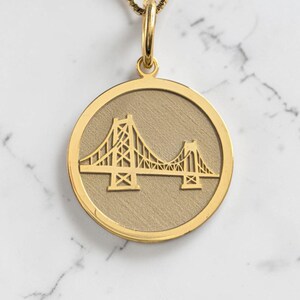 May include: A gold-colored pendant necklace featuring a detailed bridge design within a circular frame. The pendant hangs from a delicate gold chain. The bridge design is a prominent feature, showcasing intricate architectural details.
