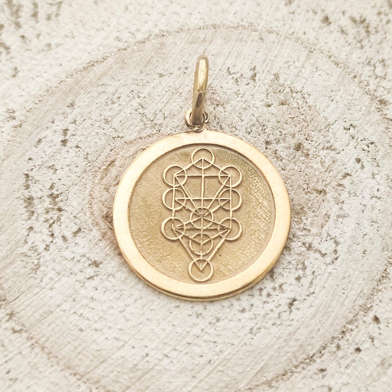 May include: A circular gold pendant featuring an engraved Tree of Life symbol. The pendant has a smooth, polished outer rim and a textured, matte-finish centre where the geometric design is etched. A small, simple gold jump ring is attached to the top.