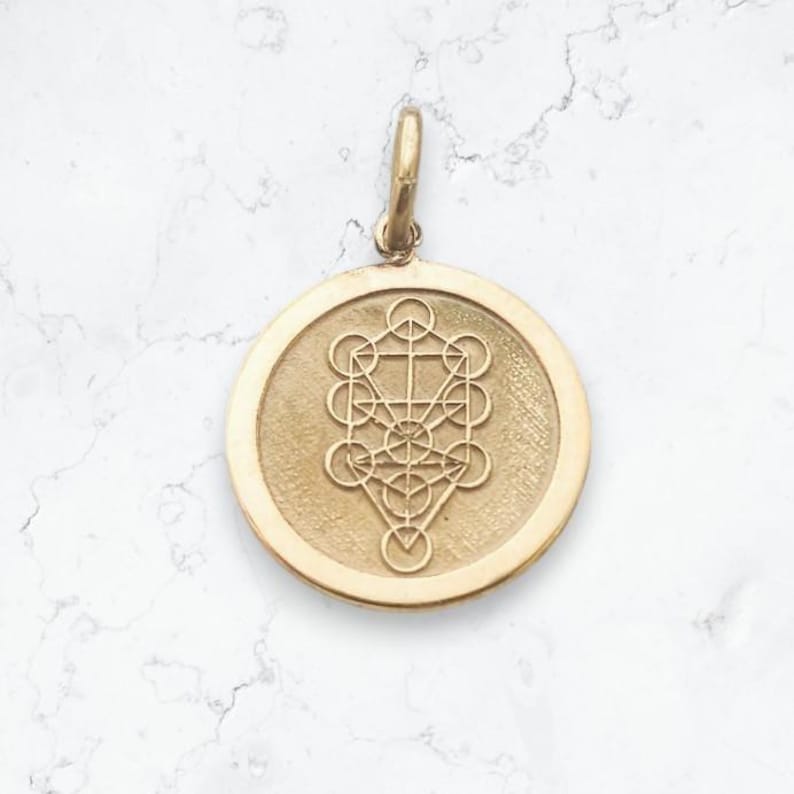 May include: A circular gold pendant featuring an engraved Tree of Life geometric design. The pendant has a smooth, raised outer rim and a textured central field. A small, vertical gold bail is attached to the top for hanging the piece.