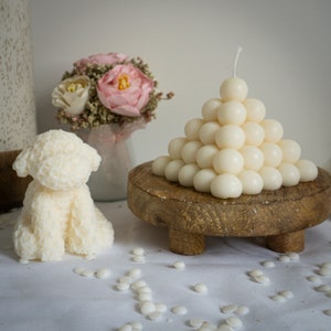 Puede incluir: Una vela decorativa con forma de caniche blanco y una vela en forma de pirámide hecha de pequeñas esferas. Las velas están sobre un soporte de madera, con un ramo de flores rosas en el fondo. Hay bolitas de cera blancas esparcidas.