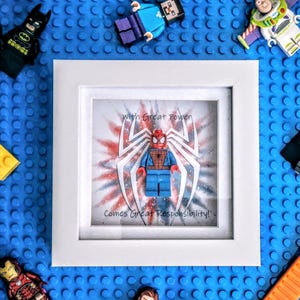 以下が含まれることがあります： 「With Great Power Comes Great Responsibility!」のテキストとスパイダーマンのフィギュアを特徴とする額入りのレゴアート作品。バットマン、ダース・ベイダー、バズ・ライトイヤーなどのレゴフィギュアが青いレゴベースに配置されています。
