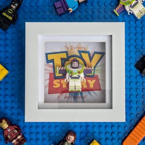 May include: A white framed Toy Story art piece featuring a Buzz Lightyear Lego figure. Surrounding the frame are various Lego figures including Batman, Darth Vader, and Iron Man, set against a blue Lego baseplate. The artwork includes the text "To Infinity and Beyond!"