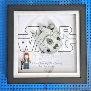 May include: Framed artwork with a Lego Millennium Falcon model and a Lego figure. The background shows the Star Wars logo and the text "A Long Time Ago In a Galaxy, Far, Far Away...". The frame is black with a white mount.