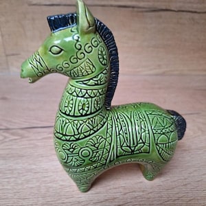 Peut inclure: Figurine de cheval en céramique verte avec des détails noirs. Le cheval présente des motifs gravés et un design stylisé. La crinière et la queue sont noires. Objet de décoration.