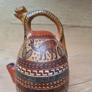 May include: A brown ceramic pitcher with a llama and cactus design. The pitcher has a unique spout and handle. The pitcher is decorated with geometric patterns in shades of brown, orange, and black.