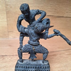 Peut inclure: Une sculpture en bronze détaillée représentant deux personnages dans une pose dynamique. Une figure est au-dessus de l'autre, avec des détails complexes sur leurs vêtements et leurs corps. La sculpture est montée sur une base rectangulaire avec un motif, mettant en valeur une esthétique vintage.