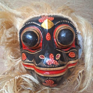 May include: A wooden mask with a black, red and gold paint design. The mask has large eyes, a wide mouth and a long, pointed nose. The mask is decorated with small, white dots. The mask is surrounded by a fringe of brown hair.
