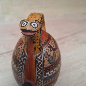 May include: A ceramic pitcher with a brown, orange, and black geometric design. The pitcher has a spout and a handle shaped like a stylized animal head with white eyes.