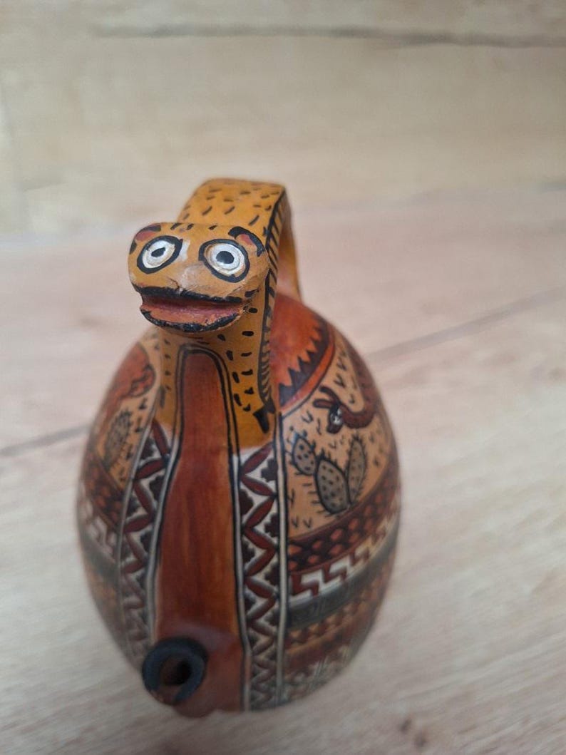 May include: A ceramic pitcher with a brown, orange, and black geometric design. The pitcher has a spout and a handle shaped like a stylized animal head with white eyes.
