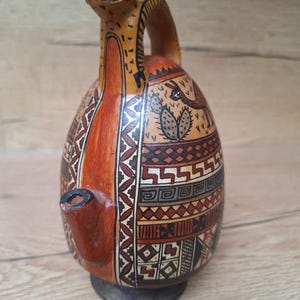 May include: A brown ceramic pitcher with a geometric pattern and a stylized animal design. The pitcher has a spout shaped like a bird's head and a handle.