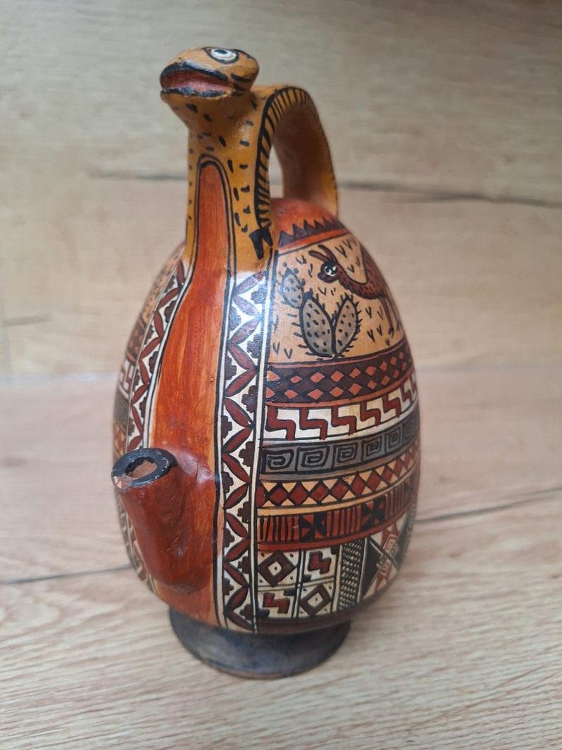 May include: A brown ceramic pitcher with a geometric pattern and a stylized animal design. The pitcher has a spout shaped like a bird's head and a handle.