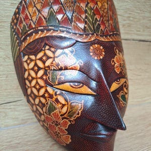Può includere: Una maschera di legno con intagli intricati e disegni batik colorati. La maschera presenta un viso umano stilizzato con occhi grandi e un naso prominente.