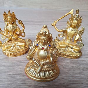 Op de afbeelding: Drie goudkleurige beelden van godheden in zittende houding. De linkerfiguur heeft de handen in een gebedsgebaar, de middelste figuur bevat kleine rode en blauwe ingelegde stenen en de rechterfiguur houdt een zwaardachtig object boven het hoofd.