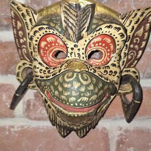 May include: A hand-carved wooden mask with a detailed design. The mask features red eyes, a green and gold face, and a black metal hanging loop. The mask has a detailed pattern and is likely used for decorative purposes.