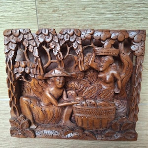 May include: A detailed, brown wooden carving depicting two figures in a natural setting. One figure wears a hat and sits near a basket, while the other carries a basket on their head. The carving features intricate details of trees and foliage.