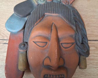 Pode incluir: Uma máscara de terracota com um rosto humano estilizado, pintado em tons de marrom, vermelho e azul. A máscara tem um cocar de penas de várias cores, incluindo verde, amarelo e branco.