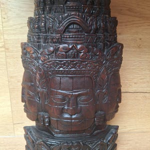 May include: A dark brown, intricately carved wooden sculpture of a multi-faced deity. The sculpture features multiple serene faces, ornate headdresses, and detailed carvings, showcasing a traditional artistic style. The wood has a rich, polished finish.