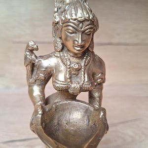 Peut inclure: Statue en laiton d'une figure féminine, ornée de bijoux complexes et d'une coiffe détaillée. La figure tient un objet en forme de bol. La statue a une teinte dorée chaude et se détache sur un fond neutre.