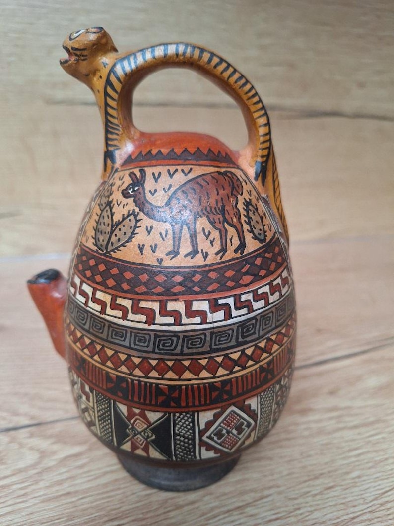 May include: A brown ceramic pitcher with a llama and cactus design. The pitcher has a unique spout and handle. The pitcher is decorated with geometric patterns in shades of brown, orange, and black.