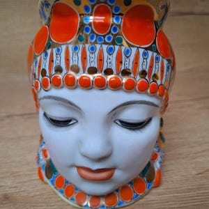 May include: A decorative ceramic vase shaped like a woman's face. The vase features a white face with closed eyes and orange lips. The headpiece is adorned with orange, blue, and gold accents. The vase is a unique piece of home decor.