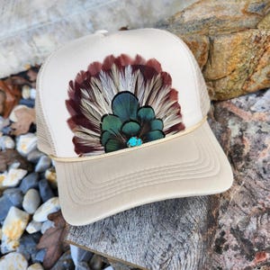 May include: A cream-coloured trucker hat with a tan brim and mesh back. The front displays a design of feathers in shades of brown, teal, and white, with a small turquoise stone at the centre. A gold-coloured rope detail accents the brim.