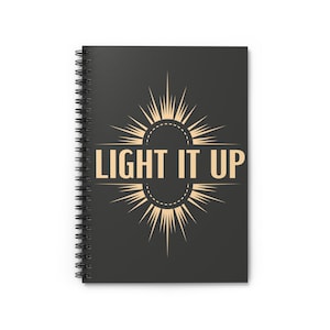 Op de afbeelding: Een zwart spiraalnotitieblok met een gouden sterburst-ontwerp en de tekst "LIGHT IT UP" in het midden.