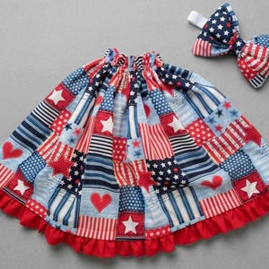 May include: A gathered skirt and a matching bow tie featuring a patchwork pattern of red, white, and blue fabric. The design includes various prints of stars, stripes, and hearts. The skirt has a solid red ruffled hem at the bottom edge.
