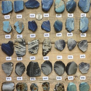 May include: A collection of 36 pieces of blue, gray, and black sea glass. The sea glass is arranged in rows on a wooden surface. Each piece of sea glass is numbered.