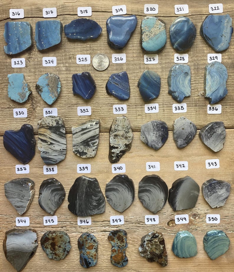 May include: A collection of 36 pieces of blue, gray, and black sea glass. The sea glass is arranged in rows on a wooden surface. Each piece of sea glass is numbered.