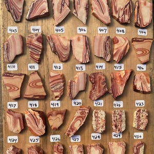 K&ouml;nnte beinhalten: Eine Sammlung von 24 einzigartigen, bunten und gemusterten Steinplatten, nummeriert von 396 bis 430, die in Reihen auf einer Holzoberfl&auml;che angeordnet sind.
