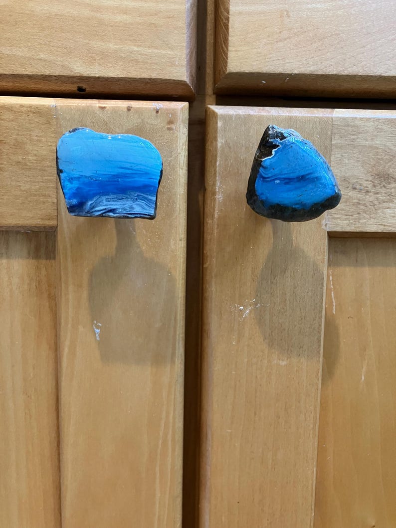 May include: Two blue and black resin cabinet knobs with a unique, abstract design. The knobs are shaped like irregular, flat rocks.