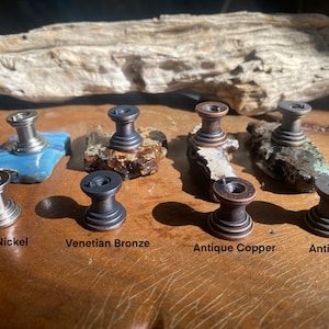 K&ouml;nnte beinhalten: Nahaufnahme von acht Schrankkn&ouml;pfen in verschiedenen Ausf&uuml;hrungen: Satin-Nickel, venezianisches Bronze, Antik-Kupfer und Antik-Messing. Die Kn&ouml;pfe werden auf einer Holzoberfl&auml;che pr&auml;sentiert.