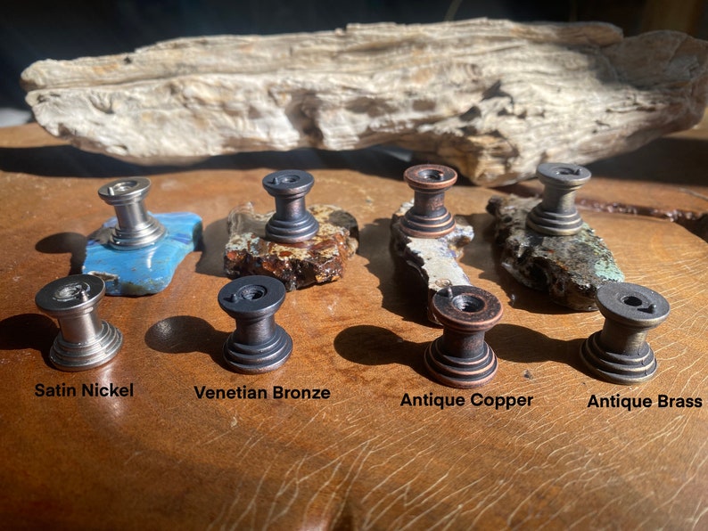 K&ouml;nnte beinhalten: Nahaufnahme von acht Schrankkn&ouml;pfen in verschiedenen Ausf&uuml;hrungen: Satin-Nickel, venezianisches Bronze, Antik-Kupfer und Antik-Messing. Die Kn&ouml;pfe werden auf einer Holzoberfl&auml;che pr&auml;sentiert.